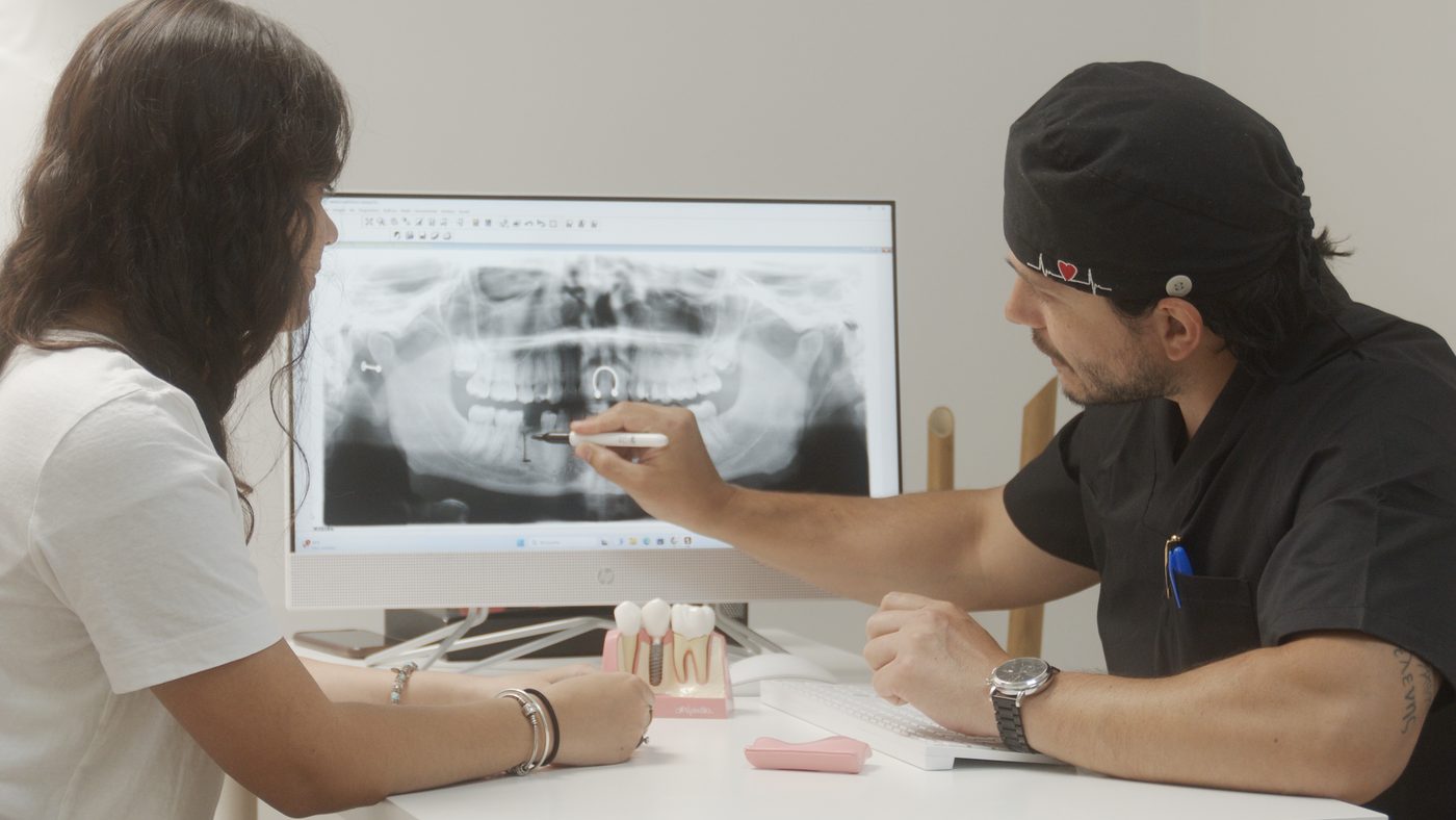 El Dr. Correa explica a una paciente su radiograf&iacute;a panor&aacute;mica en pantalla, con una maqueta de implante sobre la mesa